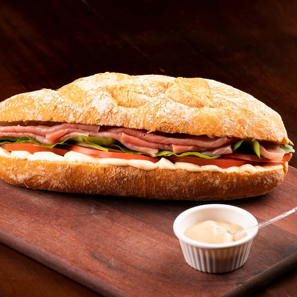 A Deli Counter Sandwich Shop sandwich with ham, lettuce, tomato, and mayonnaise on a baguette, with a side of sauce.