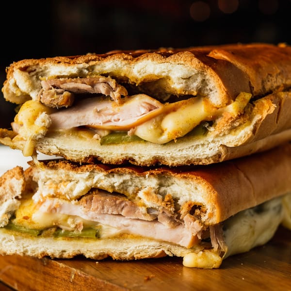A Deli Counter Sandwich Shop Cuban sandwich, cut in half, revealing melted cheese, pork, and pickles.